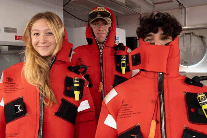 Three people wearing bright orange survival suits stand indoors on a vessel. The front person has the suit hood partially unzipped, revealing a curly-haired head. The person in the middle wears a red cap beneath the suit hood. The third individual is smiling and has long hair visible outside the suit. The environment suggests a maritime setting.