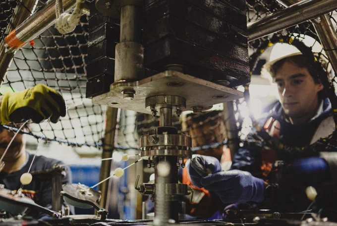 A person wearing protective gloves operates machinery surrounded by a metal framework and netting. Components and wires are visible, indicating a technical or industrial setting. The individual is focused on their task.