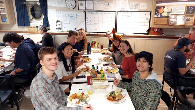A group of people seated around a table in a room with a nautical theme, enjoying a meal together. Bottles of condiments are visible on the table. The atmosphere is relaxed and casual.