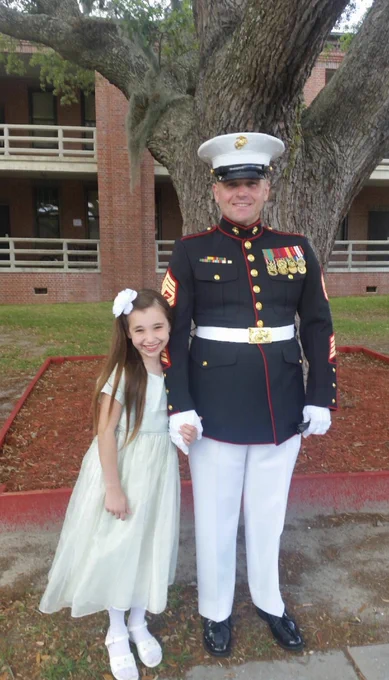 A person in a Marine Corps dress uniform stands beside a child wearing a light-colored dress with a bow in their hair. They are outdoors in front of a brick building with trees and smile at the camera.