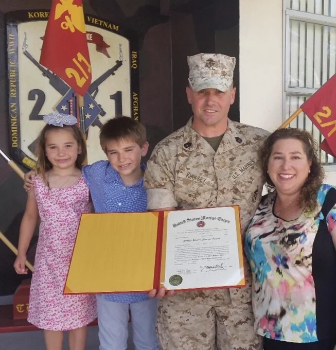A person in military uniform is holding a certificate with three people standing beside them, in front of a display. Two flags marked "2/1" are visible in the background.