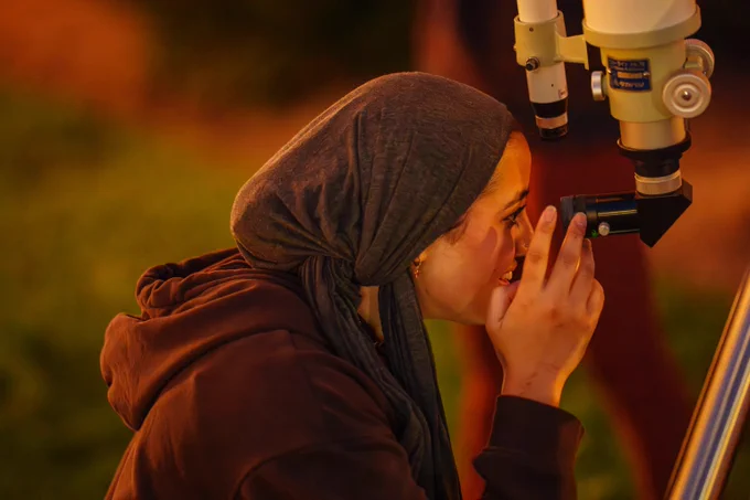 Vanderbilt student in a gray headscarf and brown hoodie observing the sky with a telescope in a natural setting.
