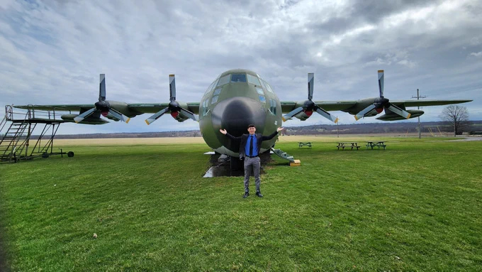 Calvin Pham stands in front of a large, green military aircraft, posing with outstretched arms. The aircraft is parked on a grass field. The sky is overcast, and picnic tables are visible in the background.