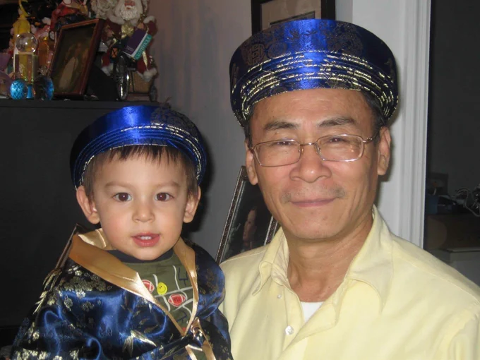 Pham Quang Khiêm and baby Calvin Pham wear matching blue hats and garments adorned with gold patterns. They are indoors with a shelf in the background displaying various decorative items.