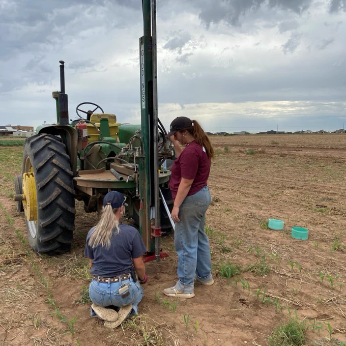 Two people are taking soil samples with heavy equipment attached to a John Deere tractor in a field. The sky is overcast, and there are houses and other structures in the distance. Some small green plants are visible in the foreground.