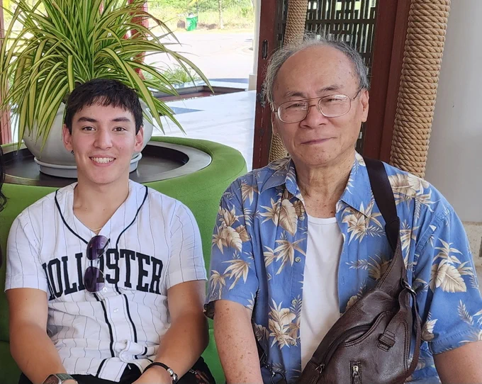 Calvin Pham, wearing a Hollister shirt, and Pham Quang Khiêm, wearing a blue floral shirt, sitting side by side, smiling at the camera. They are indoors with a plant visible in the background.