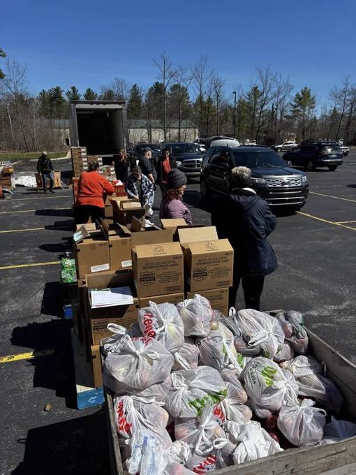 A group of people at an outdoor food distribution event in a parking lot, with packaged goods and bags of food arranged on tables. A truck is in the background, and cars are parked nearby. Trees border the area under a clear blue sky.
