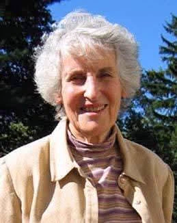 Headshot of mathematician Joan Birman, smiling and wearing a pink and white striped turtleneck with a beige jacket in an outdoor setting with blue sky and trees in the background.