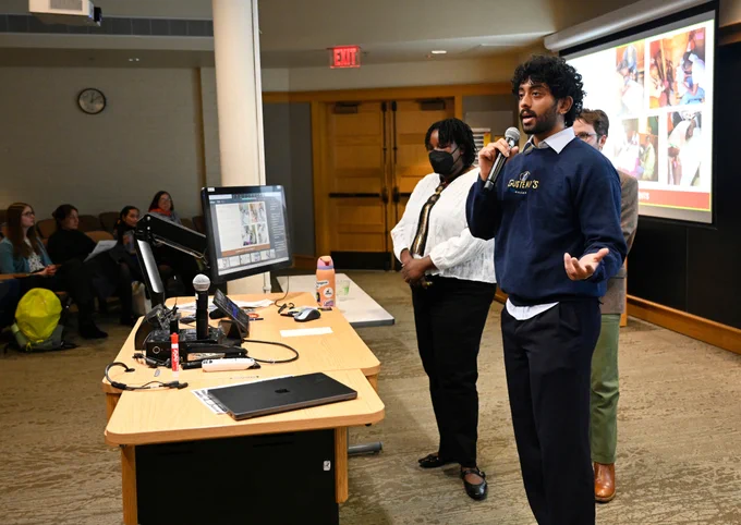 Three individuals are standing in a classroom delivering a presentation. One person is speaking into a microphone, while the other two stand nearby. A projected screen behind them displays various colorful images. The room has a few seated attendees.