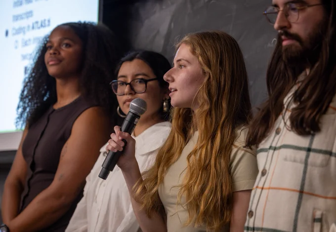 A group of four people stand in front of a chalkboard. One person is speaking into a microphone. 
