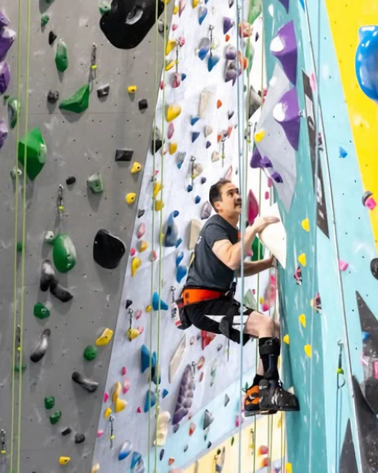 A person with a prosthetic leg climbs an indoor rock wall with colorful holds, wearing a harness and climbing gear.