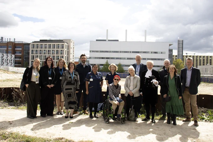 A group of thirteen people, including a wheelchair user, standing together outdoors in front of the site of Cambridge Cancer Research Hospital