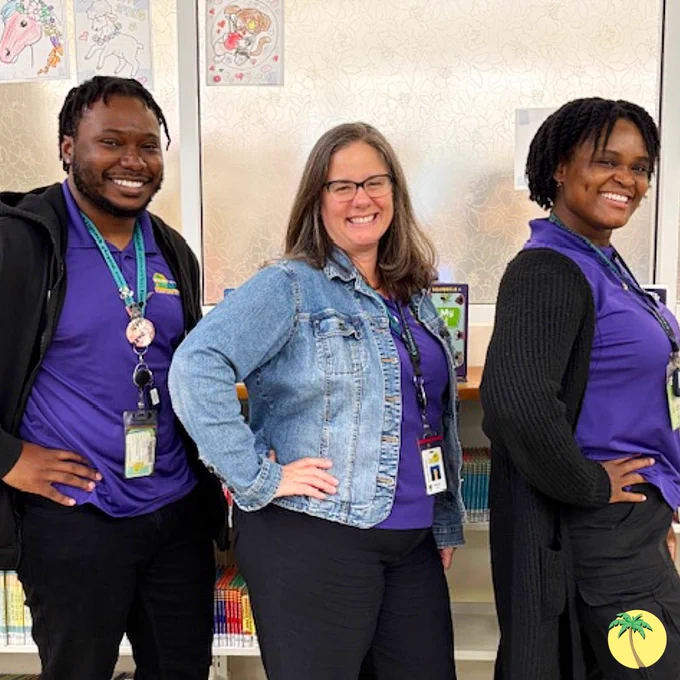 Three Library staff in matching purple Library shirts, posing with their hand on their hip and smiling at the camera.
