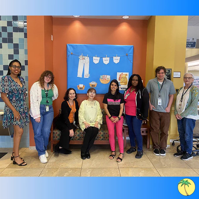 Library staff gathered around a sofa, all dressed colorfully. From left-to-right we have a woman in a multi-colored dress, a woman with a bright green shirt, a woman with a bright orange shirt, a woman with a pastel yellow shirt, a woman with hot pink pants, a woman with a pink Library shirt, a man with a gray Library shirt, and a woman with pastel green cardigan.