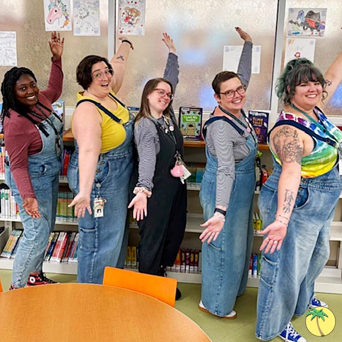 Staff posing with their arms diagonal like showgirls, smiling at the viewer. The five staff are in overalls with brightly colored shirts: from left-to-right, they're in red, yellow, gray, gray, and tie-dye.