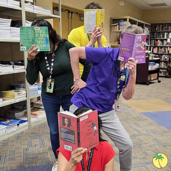 Four Library staff in four different colors: Green, yellow, purple, and red. They each hold a book in front of their face, and each book matches the color of the holder's shirt.