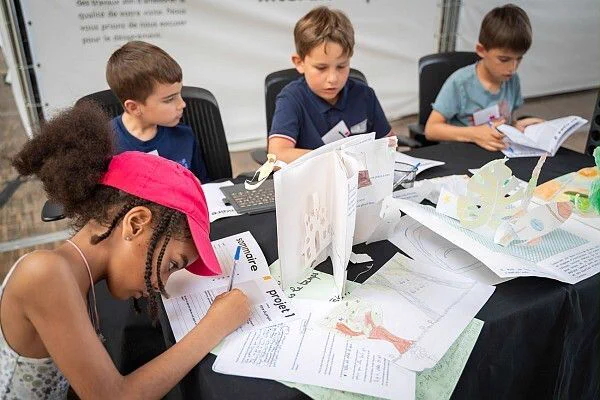 Des enfants travaillent ensemble autour d’une table, en train de dessiner, découper et assembler des maquettes, dans une démarche créative et collaborative. 