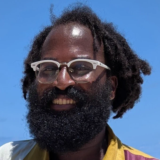 Mugabi Byenkya a dark skinned person with glasses with white frames, black free form locs and a luscious full black beard. Smiling. 