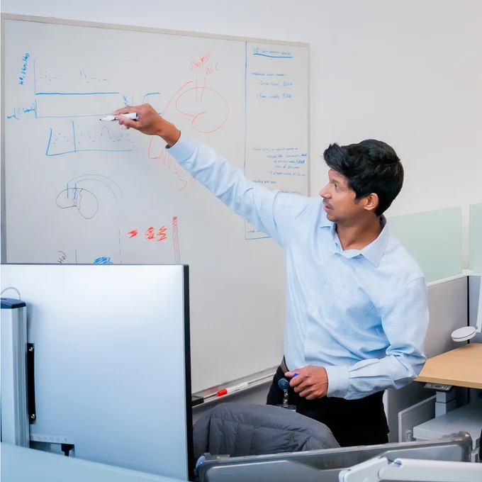 Man with short black hair wearing a blue shirt standing and drawing on a white board with diagrams in blue and red.