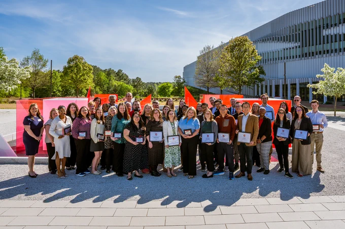 A group of staff and faculty hold certificates in front of the Reds and Whites outside Hunt Library.