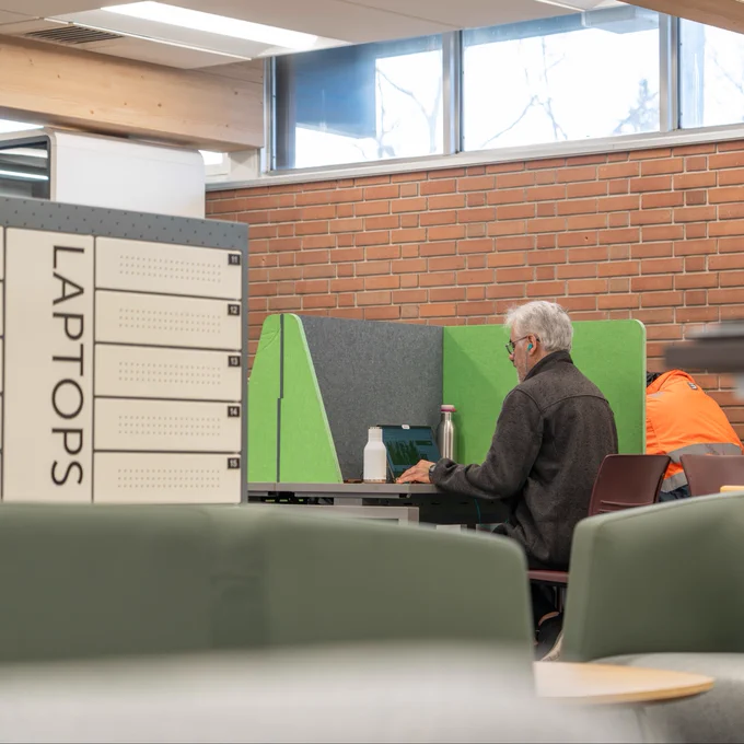 A person sits at a library desk with a green privacy screen, using a laptop. A cabinet with the word "LAPTOPS" is visible on the left. 