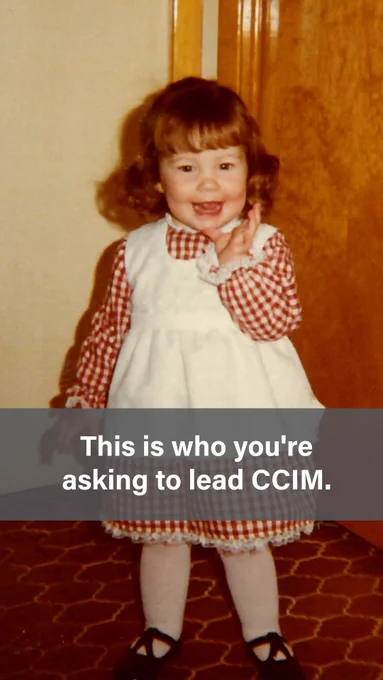 A vintage childhood photo of a smiling toddler girl with short curly reddish-brown hair, wearing a red and white gingham long-sleeve shirt under a white pinafore dress with lace trim, standing in front of a wooden door on a patterned carpet. Text overlay reads: "This is who you're asking to lead CCIM."
