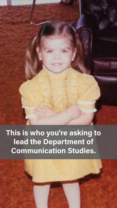 A vintage childhood photo of a toddler girl with light brown hair in two small pigtails with pink barrettes, wearing a yellow short-sleeve knit dress with floral details and white trim, standing with her arms crossed in front of a dark leather armchair on a red-orange carpet. Text overlay reads: "This is who you're asking to lead the Department of Communication Studies."