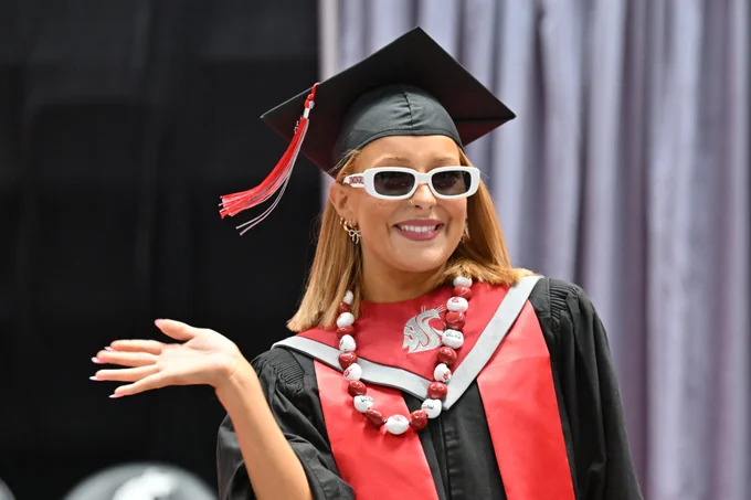 Person standing on a graduation stage wearing a black mortarboard cap with a red tassel, a black academic gown, and a wide red stole. A gray emblem is visible on the stole, and a red and white beaded necklace with printed text hangs around the neck. The person wears white, rectangular sunglasses, small hoop earrings, and has shoulder length light brown hair visible beneath the cap. One arm is bent at the elbow with the palm facing up in a presenting gesture. The background consists of softly lit, vertical gray stage curtains, with a dark, out of focus area to the left indicating the stage setting.