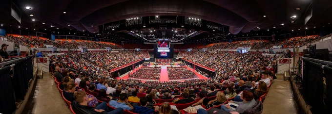 Wide panoramic view inside a large indoor arena during a university commencement ceremony, with rows of graduates seated on the floor around a central red aisle and a stage at the far end. The surrounding stadium seating is filled with spectators on multiple levels. Overhead lighting illuminates the arena, and large video screens above the stage display the ceremony proceedings.