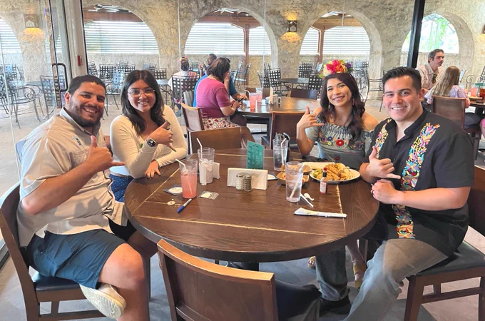 A group of four TAMU-CC Islander alumni sit around a dining table at a restaurant. They are smiling, wearing festive Mexican traditional clothing, and giving shakas-up gestures. The table has drinks and plates of food. The restaurant has stone walls and large windows in the background.