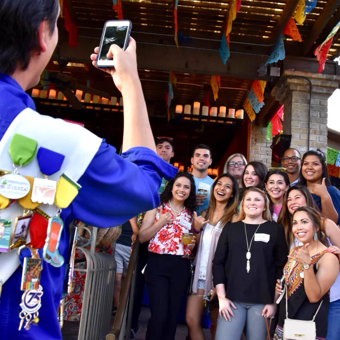 A group of TAMU-CC Islander alumni are posing for a photo at an outdoor gathering, with colorful papel picado decorations hanging above. The person taking the photo has a blue shirt adorned with various festive pins and badges on the back.