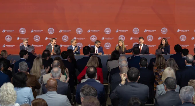 A panel sits at a table with an orange backdrop displaying \"Michael & Susan Dell FOUNDATION\" and \"UNIVERSITY OF TEXAS AT AUSTIN\" logos. The audience applauds. The setting suggests a press conference or formal announcement.