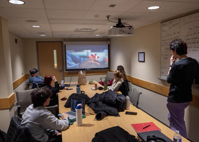 A group of people are sitting and standing around a conference table in a meeting room, watching a projection screen displaying an image. The room has a whiteboard with writing on it, a door, and various items like notebooks, water bottles and coats on the table.