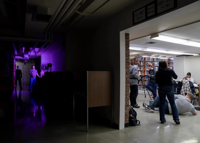 On the left is a dimly lit room with a person standing with a purple light casting shadows. On the right, a regularly-lit room hosts a group of people sitting or stand around tables and bookshelves in Alden Library.