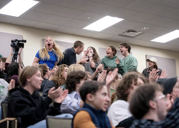 Six winners from the champion team of the 48-Hour Shootout competition stand up from their seats in a lecture hall to celebrate the announcement of their win. The remaining people in the room are seated and many are clapping. 