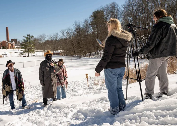 A group of people stands in a snowy landscape. Three individuals wearing winter clothing are on the left, one with a colorful cloth poncho. On the right, two people are operating a camera on a tripod. Trees and a red brick chimney are visible in the background under a clear blue sky.