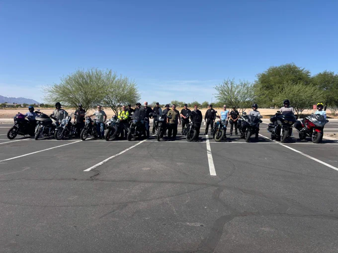Group of motorcyclists lined up with their bikes in a sunny parking lot under clear blue sky.