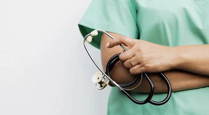 A person wearing a green medical scrub top is holding a stethoscope in their crossed arms.
