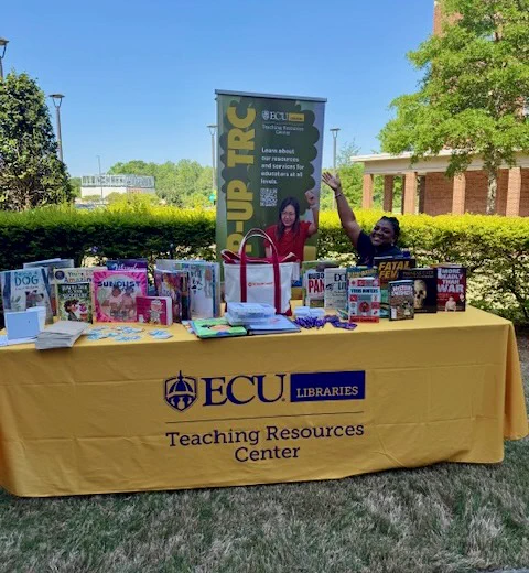 A person sitting behind a table covered with various books and materials. The tablecloth displays the "ECU Libraries Teaching Resources Center" logo. The scene is outdoors with a building and trees in the background.