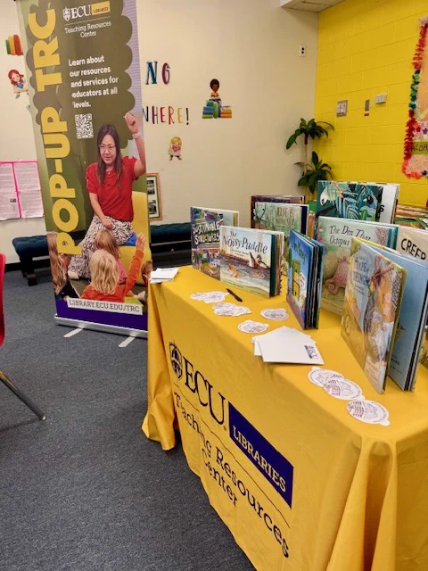 A library display features books set up on a table with a bright yellow tablecloth, which has the "ECU Libraries Teaching Resources Center" logo. To the left, a pop-up banner with text about ECU's teaching resources is visible. The room has colorful walls and decorations, giving it a cheerful atmosphere.