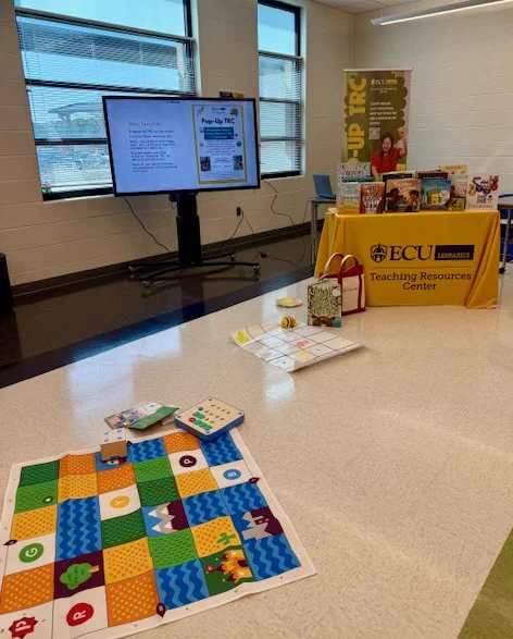 A classroom setup with a display area featuring an ECU Libraries Teaching Resources Center table covered with educational materials and books. A large screen shows information. Educational games and mats are spread on the floor, and bright windows illuminate the space.