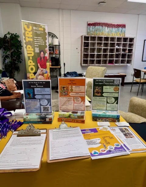 This image shows a display table with informational brochures and clipboards on a yellow tablecloth. Behind the table, there's a pop-up banner featuring promotional content. Comfortable chairs and a wall of cubby holes are visible in the background.