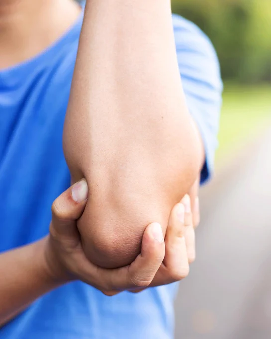 A person in a blue shirt holding their elbow with one hand, outdoors in a blurred natural setting.