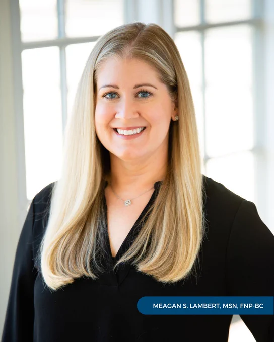 Meagan S. Lambert, MSN, FNP-BC, of Tryon Medical Partners smiles and wears a black blouse standing in front of a paned glass window