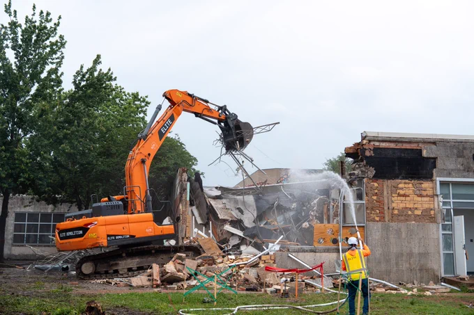 An orange excavator demolishing a building with a worker in a safety vest nearby.