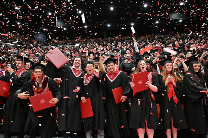 Large group of graduates seated and standing inside a packed indoor arena during a commencement ceremony, wearing black caps and gowns with red stoles and holding red diploma covers as red and white confetti falls from above. The floor seating is filled with graduates, the surrounding stadium seats are crowded with spectators, and bright overhead lights illuminate the celebration.