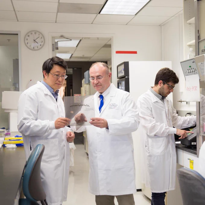 Three researchers in lab coats work in a laboratory. Two are examining a transparent sheet with data, while the third operates equipment at a bench. A clock and various lab instruments are visible in the room.
