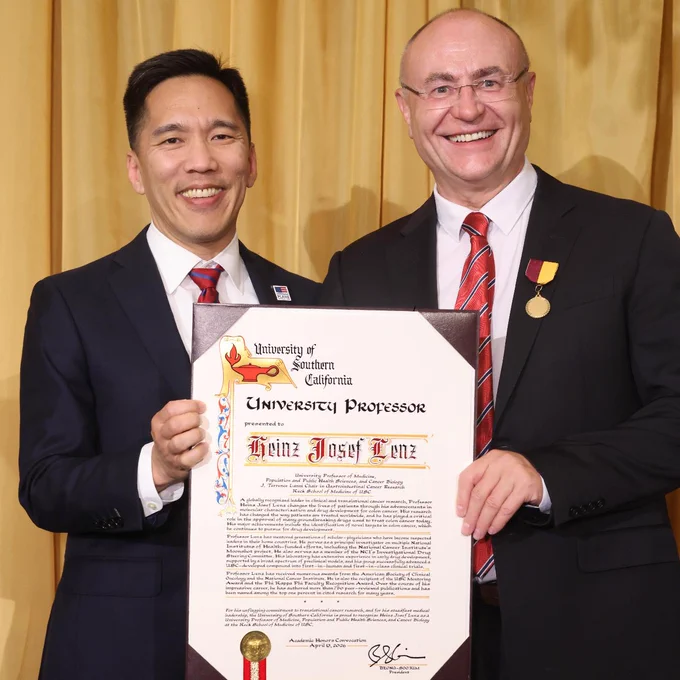 Two individuals in formal attire are holding a framed document from the University of Southern California, which names Heinz Josef Lenz as a University Professor. One individual is wearing a medal. They are standing in front of a gold curtain.