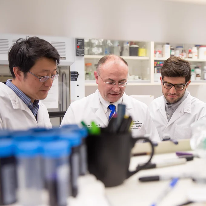 A group of three people in a laboratory setting, wearing white lab coats, engage in discussion around a table with scientific equipment and containers. The background shows shelves stocked with various lab supplies.