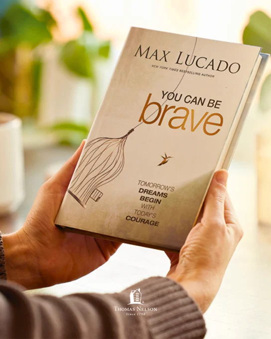 A person holds a book titled "You Can Be Brave" by Max Lucado. The cover features an illustration of a bird near a cage and includes the text "Tomorrow's dreams begin with today's courage." The publisher's logo, "Thomas Nelson," is visible at the bottom.
